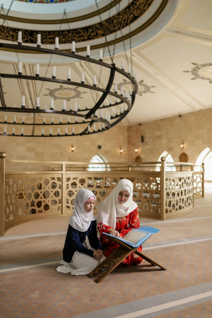 A woman and young girl in hijabs read a Quran together inside a mosque.
